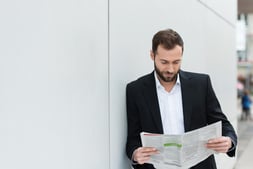 Businessman reading a newspaper on his lunch break as he leans against a white wall with copyspace.jpeg