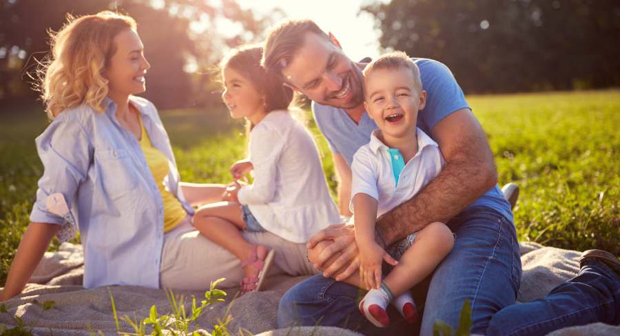 Happy family with mom and dad and two kids sitting on grass in sunlight.